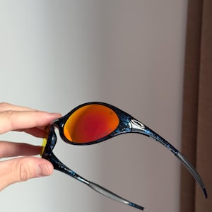 Puede incluir: Gafas de sol negras y azules con lentes naranjas. Las monturas tienen un dise&ntilde;o estampado. Las patillas son plateadas y se curvan para adaptarse a las orejas. Las lentes son de color naranja brillante.