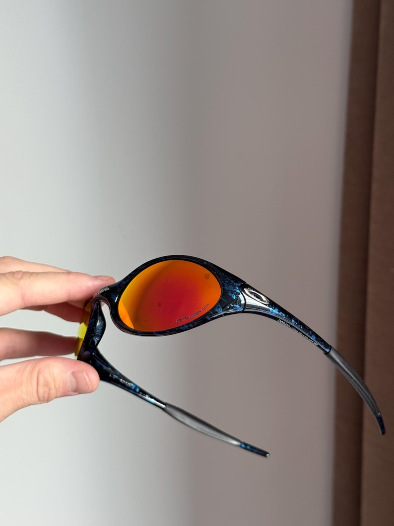 Puede incluir: Gafas de sol negras y azules con lentes naranjas. Las monturas tienen un dise&ntilde;o estampado. Las patillas son plateadas y se curvan para adaptarse a las orejas. Las lentes son de color naranja brillante.