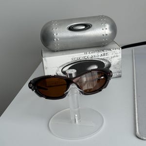 May include: Brown sunglasses with a matching case and a box with the text "SCIENCE AND ART". The sunglasses are displayed on a clear stand. The case is silver and cylindrical.