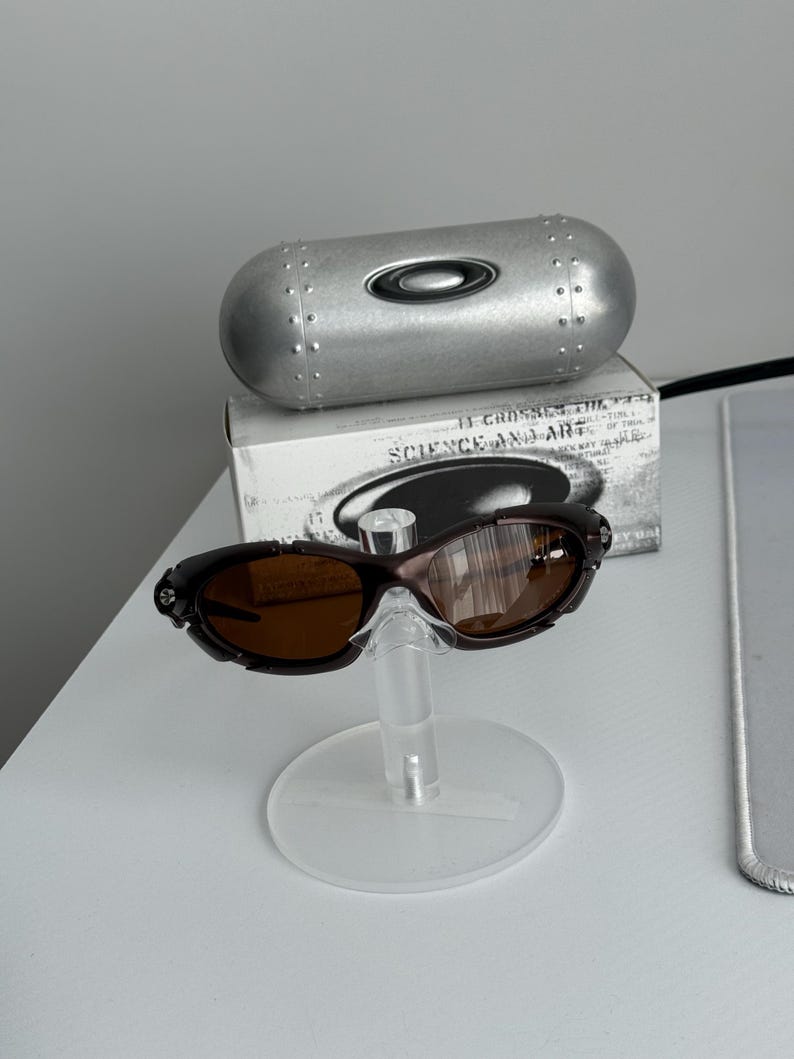 May include: Brown sunglasses with a matching case and a box with the text "SCIENCE AND ART". The sunglasses are displayed on a clear stand. The case is silver and cylindrical.
