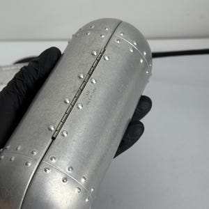May include: A silver-colored, riveted case with a hinged opening. The case has a cylindrical shape with rounded ends and is held by a gloved hand. The text "CASE MADE IN CHINA" is visible on the hinge.