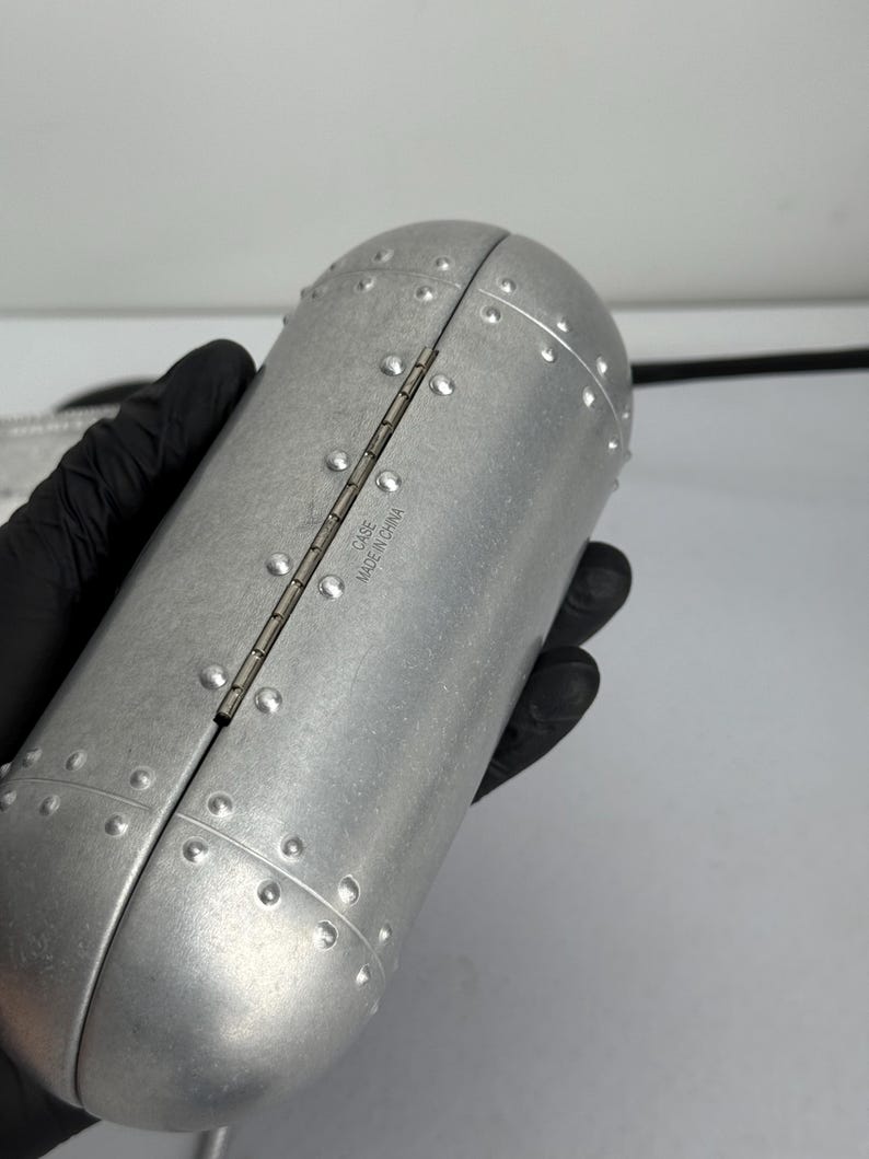 May include: A silver-colored, riveted case with a hinged opening. The case has a cylindrical shape with rounded ends and is held by a gloved hand. The text "CASE MADE IN CHINA" is visible on the hinge.