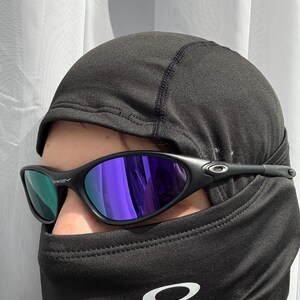 May include: Black sunglasses with purple mirrored lenses. The sunglasses have a sleek, modern design with a black frame. The image also shows a black balaclava with a white logo.
