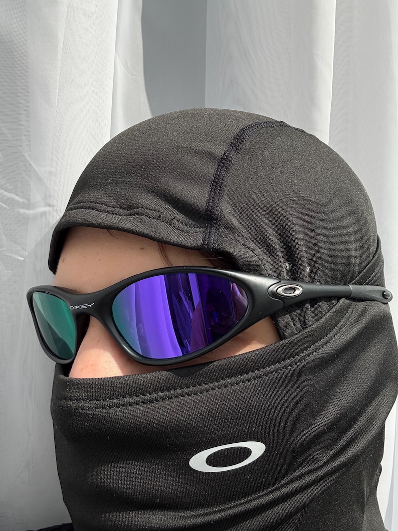 May include: Black sunglasses with purple mirrored lenses. The sunglasses have a sleek, modern design with a black frame. The image also shows a black balaclava with a white logo.