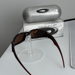 May include: Brown-framed sunglasses with brown lenses are displayed on a clear stand. A silver, cylindrical case with the Oakley logo is behind the sunglasses. A box with the text "SCIENCE AND ART" is also visible.
