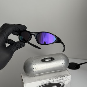 May include: Black Oakley sunglasses with purple mirrored lenses. The sunglasses are held above a silver case and a white box with the text "SCIENCE AND ART". The case has a silver finish with a circular Oakley logo.
