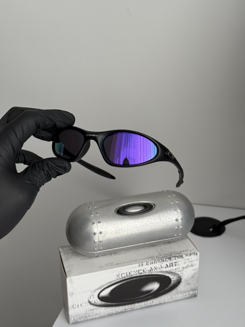 May include: Black Oakley sunglasses with purple mirrored lenses. The sunglasses are held above a silver case and a white box with the text "SCIENCE AND ART". The case has a silver finish with a circular Oakley logo.