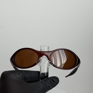 May include: Maroon-colored Oakley sunglasses with brown lenses. The sunglasses have a curved frame and a small bridge. The lenses are oval-shaped. The sunglasses are held up by a clear stand.