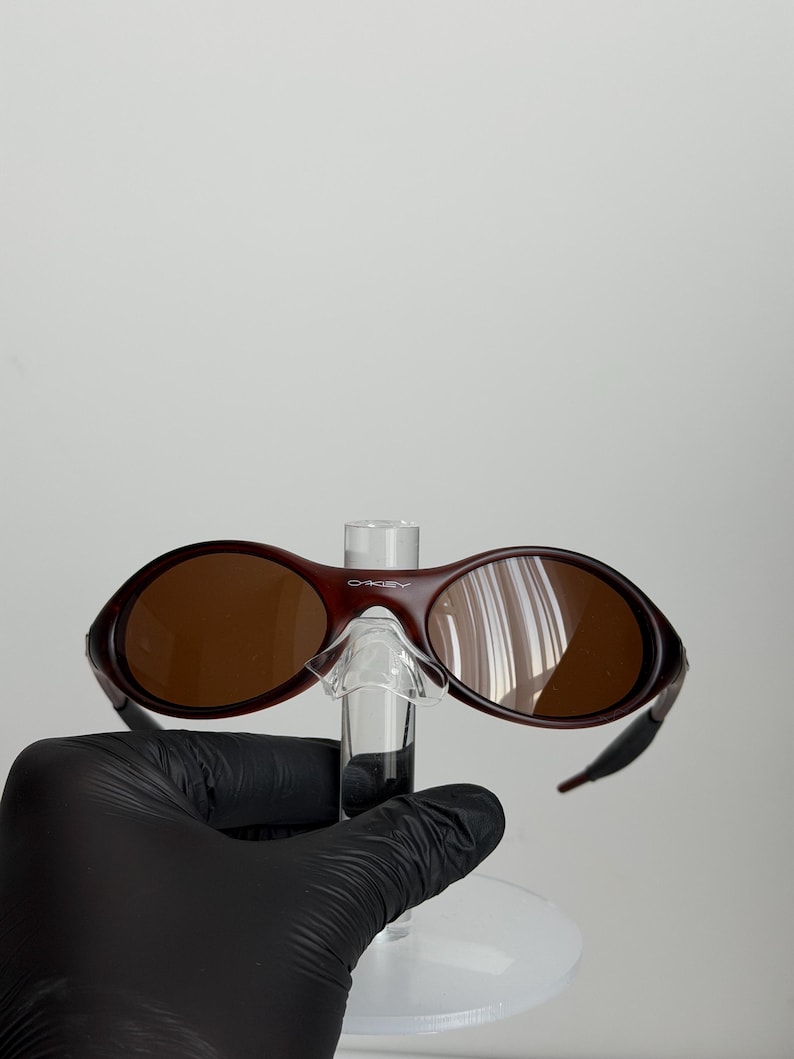 May include: Maroon-colored Oakley sunglasses with brown lenses. The sunglasses have a curved frame and a small bridge. The lenses are oval-shaped. The sunglasses are held up by a clear stand.