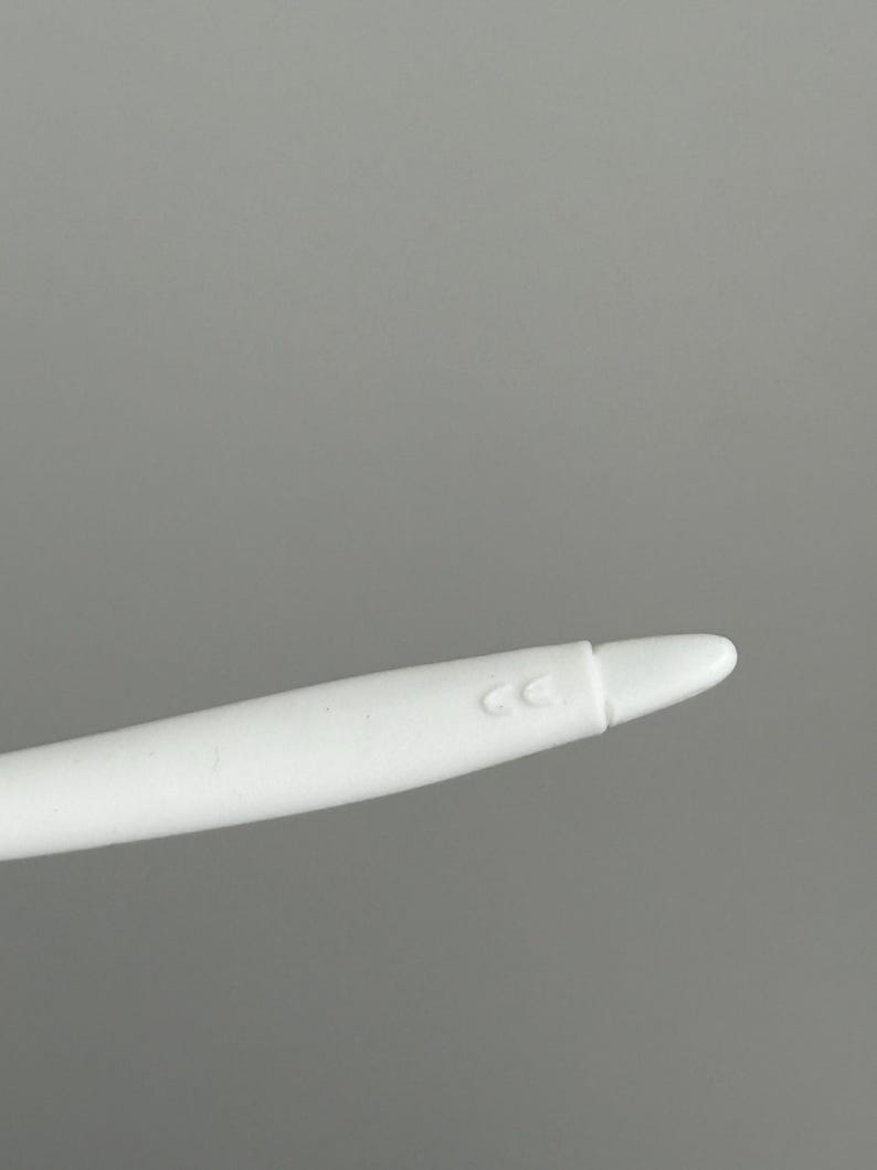 May include: A white stylus with a rounded tip. The stylus has a smooth, matte finish and a slight curve. The letters "CC" are printed on the side of the stylus.