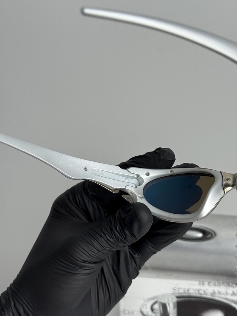 May include: Silver-framed sunglasses with blue lenses. The sleek, modern design features a metallic finish and curved temples. The sunglasses are held by a gloved hand, suggesting careful handling. The lenses appear to be tinted, offering protection from the sun.