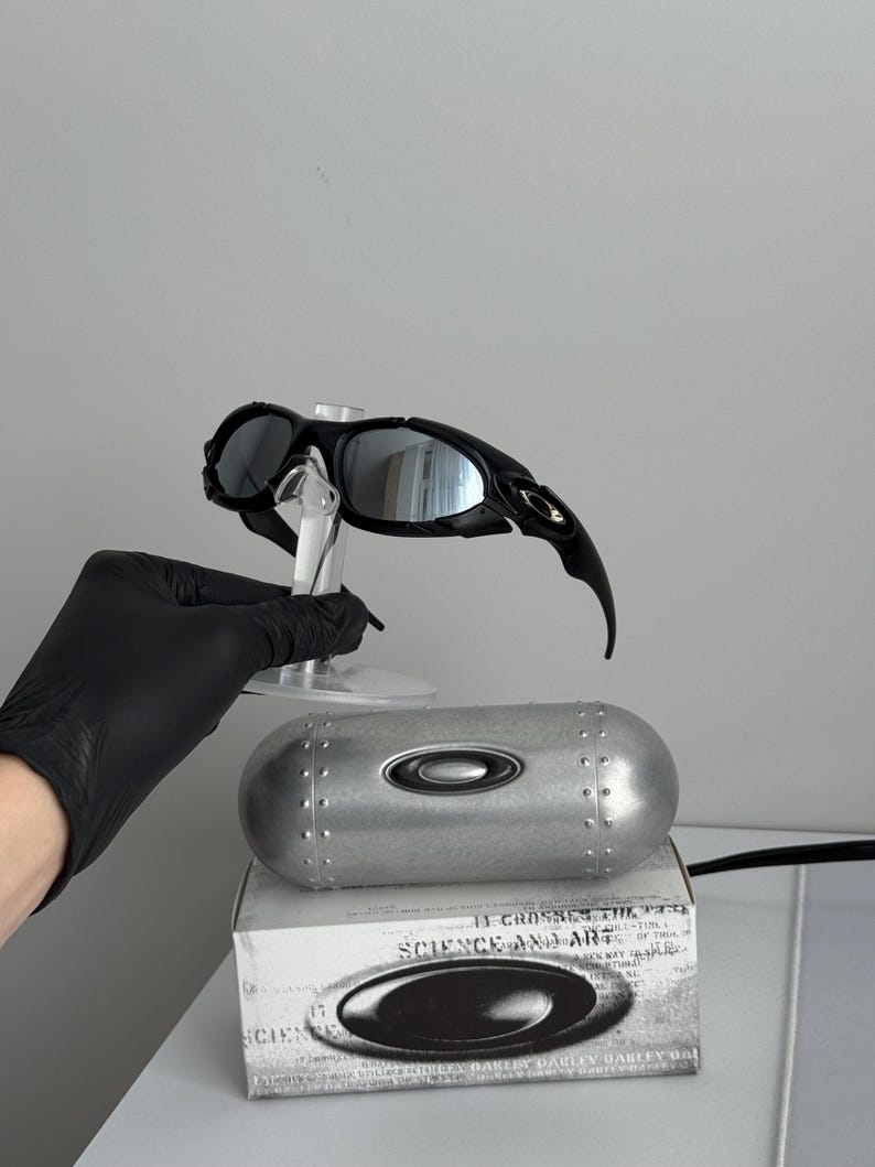May include: Black sunglasses with reflective lenses are displayed on a clear stand. A silver case and a white box with the Oakley logo and text are also visible. The sunglasses have a sleek, modern design.