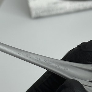 Pu&ograve; includere: Primo piano di un oggetto in metallo argentato con la parola "jacket" incisa sulla superficie. L'oggetto &egrave; tenuto da una persona che indossa un guanto nero. Lo sfondo &egrave; sfocato.