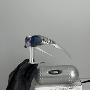 May include: Silver and blue Oakley sunglasses with a futuristic design, displayed on a clear stand. The image also shows a silver case and a white box with the text "SCIENCE AND ART".