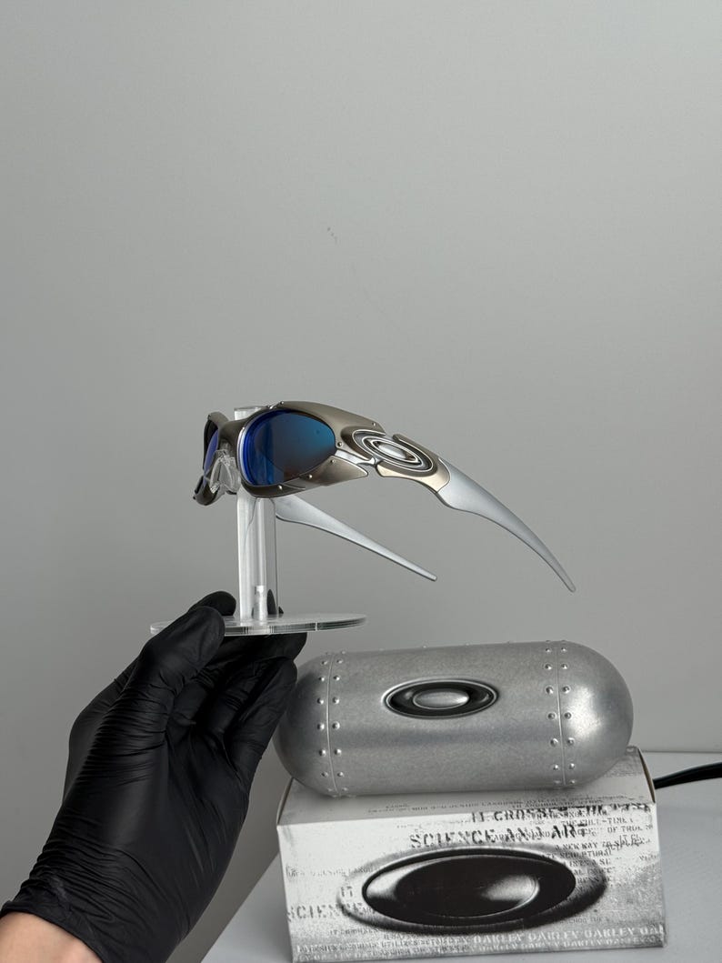 May include: Silver and blue Oakley sunglasses with a futuristic design, displayed on a clear stand. The image also shows a silver case and a white box with the text "SCIENCE AND ART".
