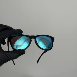 May include: Black sunglasses with blue mirrored lenses. The sunglasses have a classic frame design and are held by a gloved hand. The lenses reflect a blue hue, and the overall style is modern and sleek.