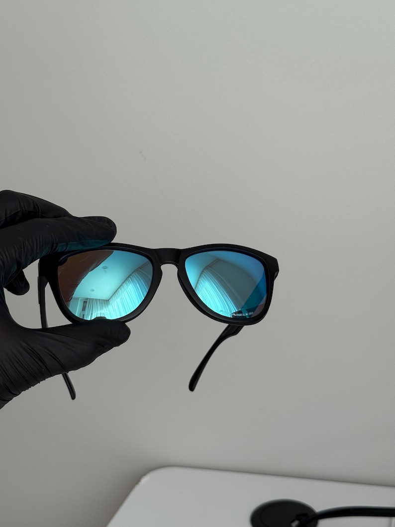 May include: Black sunglasses with blue mirrored lenses. The sunglasses have a classic frame design and are held by a gloved hand. The lenses reflect a blue hue, and the overall style is modern and sleek.