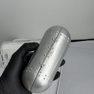 May include: A silver-colored, oval-shaped case with a hinged lid. The case is covered in small, evenly spaced rivets, giving it a metallic appearance. The text "CASE MADE IN CHINA" is visible on the hinge.