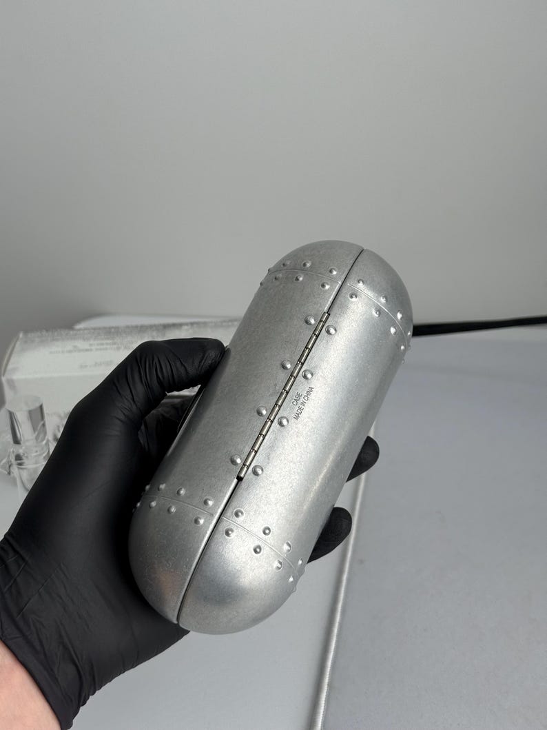 May include: A silver-colored, oval-shaped case with a hinged lid. The case is covered in small, evenly spaced rivets, giving it a metallic appearance. The text "CASE MADE IN CHINA" is visible on the hinge.