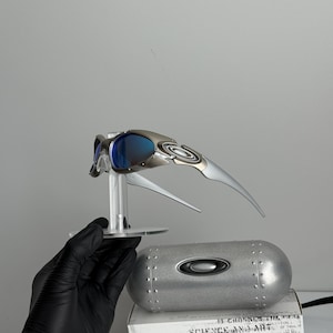 May include: Silver and blue Oakley sunglasses with a futuristic design, displayed on a clear stand. A silver case and a white box with the text "SCIENCE AND ART" are also visible.