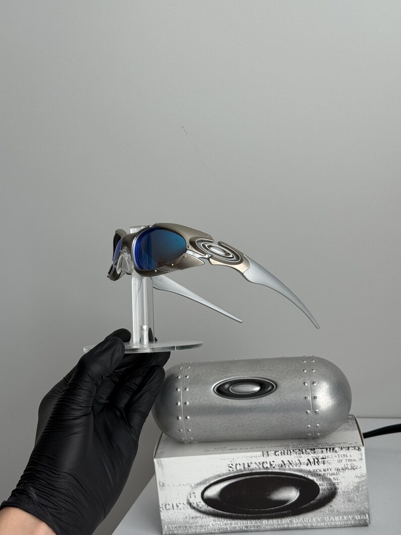 May include: Silver and blue Oakley sunglasses with a futuristic design, displayed on a clear stand. A silver case and a white box with the text "SCIENCE AND ART" are also visible.