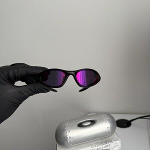 May include: Black sunglasses with purple-pink lenses are held up in front of a silver case and a box with the text "SCIENCE AND ART". The sunglasses have a sleek, modern design.