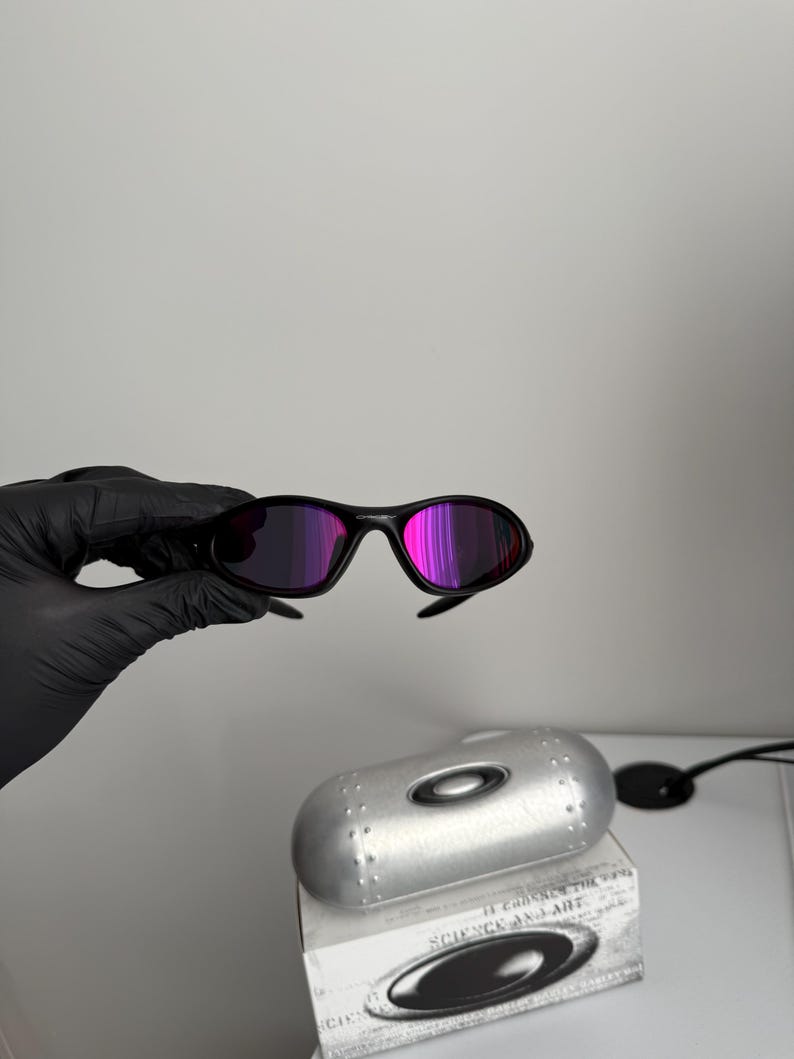 May include: Black sunglasses with purple-pink lenses are held up in front of a silver case and a box with the text "SCIENCE AND ART". The sunglasses have a sleek, modern design.
