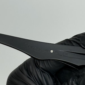 May include: Close-up of a black, plastic tool with a small, silver accent. The tool has text etched into the surface. The tool is held by a gloved hand.