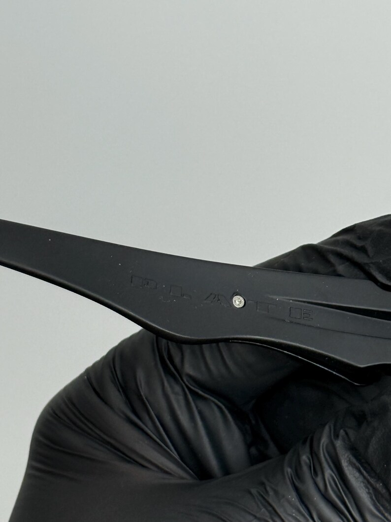 May include: Close-up of a black, plastic tool with a small, silver accent. The tool has text etched into the surface. The tool is held by a gloved hand.