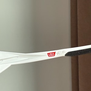 以下が含まれることがあります： 黒いテンプルが付いた白いサングラス。フレームには「USA Assembled」の文字が入った赤い長方形があります。サングラスはニュートラルな背景に配置されています。