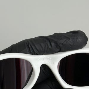 May include: White sunglasses with dark lenses. The sunglasses have a thick, rounded frame. The text "BRUTE" is visible on the top of the frame. The sunglasses are held by a gloved hand.