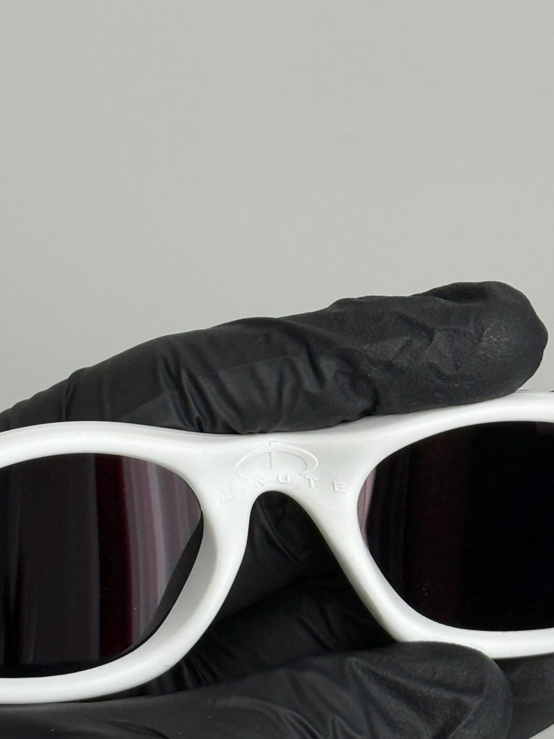 May include: White sunglasses with dark lenses. The sunglasses have a thick, rounded frame. The text "BRUTE" is visible on the top of the frame. The sunglasses are held by a gloved hand.