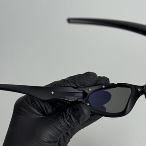 May include: Black sunglasses with a rectangular frame and dark lenses. The sunglasses have a sleek, modern design with a matte finish. The temples are thin and curve around the ears. The lenses have a slight blue tint.