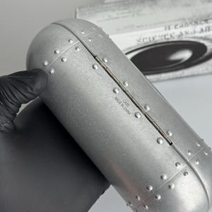 May include: A silver-colored, riveted metal case with a rounded rectangular shape. The case is hinged and has the text "MADE IN CHINA" printed on it. The background includes a partially visible box with the words "SCIENCE AND ART".