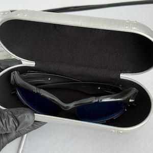 May include: Black sunglasses with blue lenses rest inside a silver case with a black interior. The sunglasses have a sleek, modern design. The case is oval-shaped and has a textured surface.