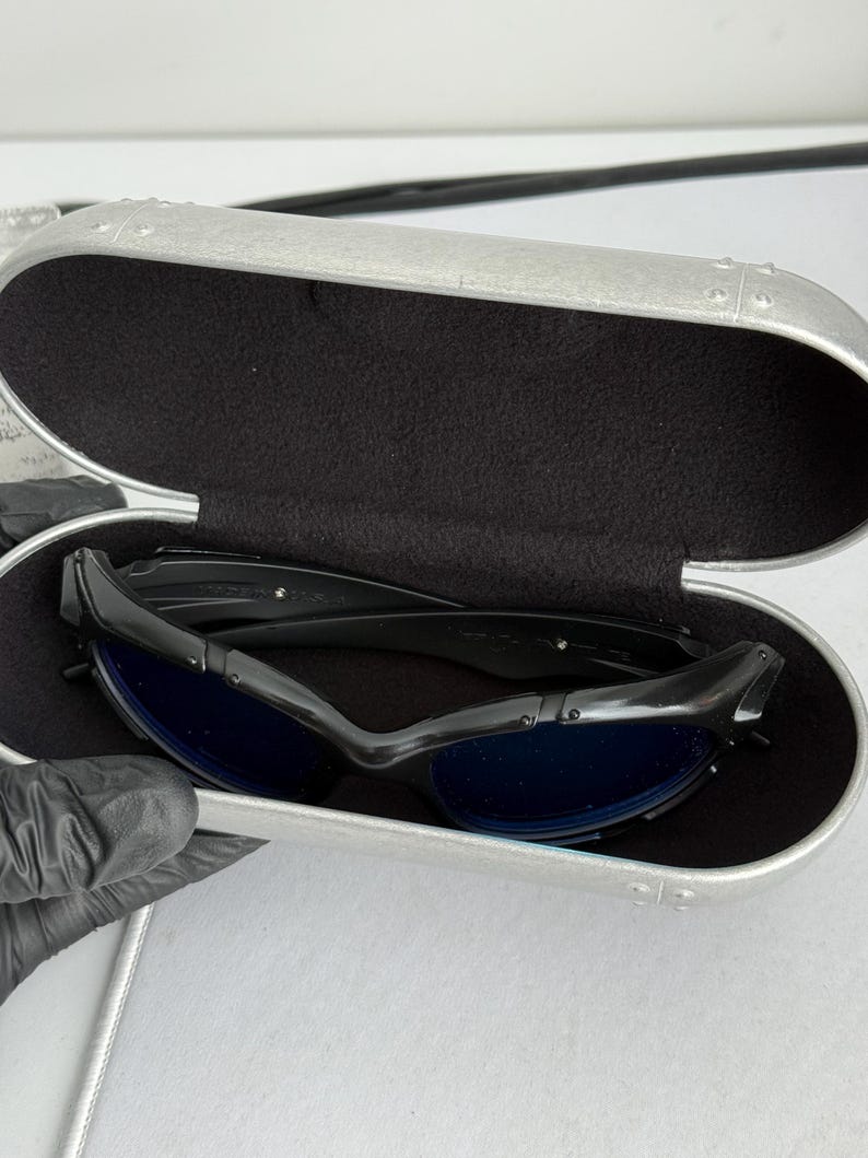 May include: Black sunglasses with blue lenses rest inside a silver case with a black interior. The sunglasses have a sleek, modern design. The case is oval-shaped and has a textured surface.