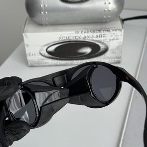 K&ouml;nnte beinhalten: Schwarze Sonnenbrille mit dunkelgrauen Gl&auml;sern. Die Sonnenbrille wird vor einer wei&szlig;en Oberfl&auml;che gehalten. Der Text "SCIENCE AND ART" ist auf der Schachtel hinter der Sonnenbrille zu sehen. Ein silbernes Etui ist ebenfalls sichtbar.