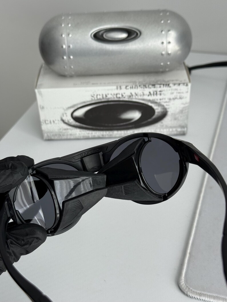 K&ouml;nnte beinhalten: Schwarze Sonnenbrille mit dunkelgrauen Gl&auml;sern. Die Sonnenbrille wird vor einer wei&szlig;en Oberfl&auml;che gehalten. Der Text "SCIENCE AND ART" ist auf der Schachtel hinter der Sonnenbrille zu sehen. Ein silbernes Etui ist ebenfalls sichtbar.