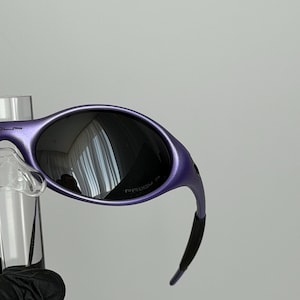 May include: Purple sunglasses with dark lenses. The frames have a curved design and a matte finish. The lenses reflect the surrounding environment. The sunglasses are displayed on a clear stand.