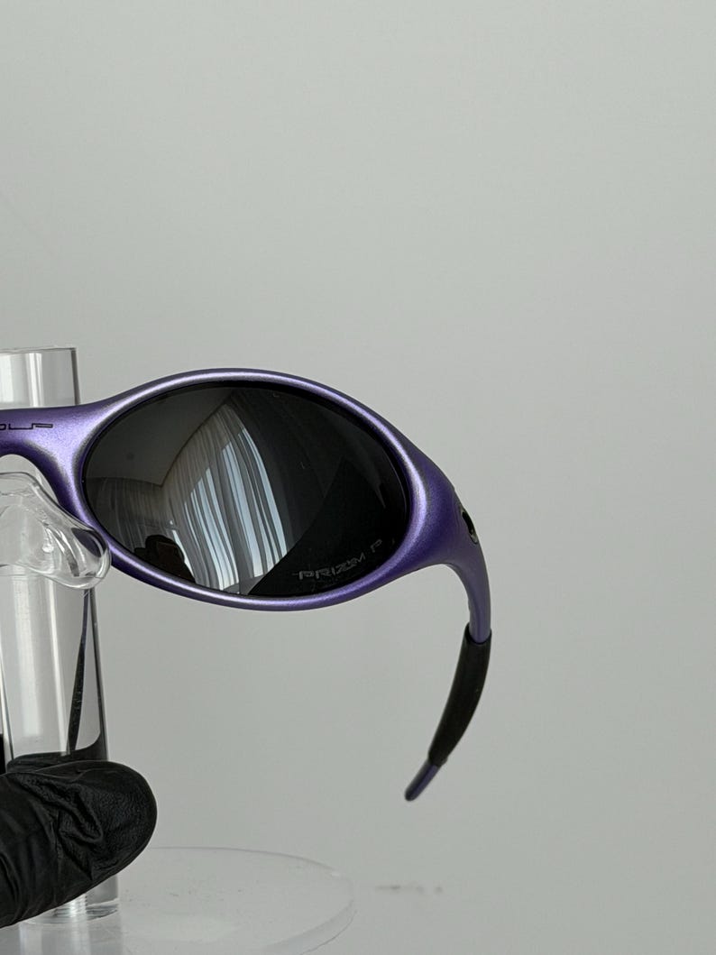 May include: Purple sunglasses with dark lenses. The frames have a curved design and a matte finish. The lenses reflect the surrounding environment. The sunglasses are displayed on a clear stand.
