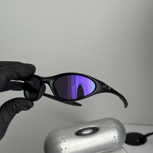 May include: Black Oakley sunglasses with purple mirrored lenses. The sunglasses are held by a gloved hand. A silver case and a box with the text "SCIENCE AND ART" are in the background.