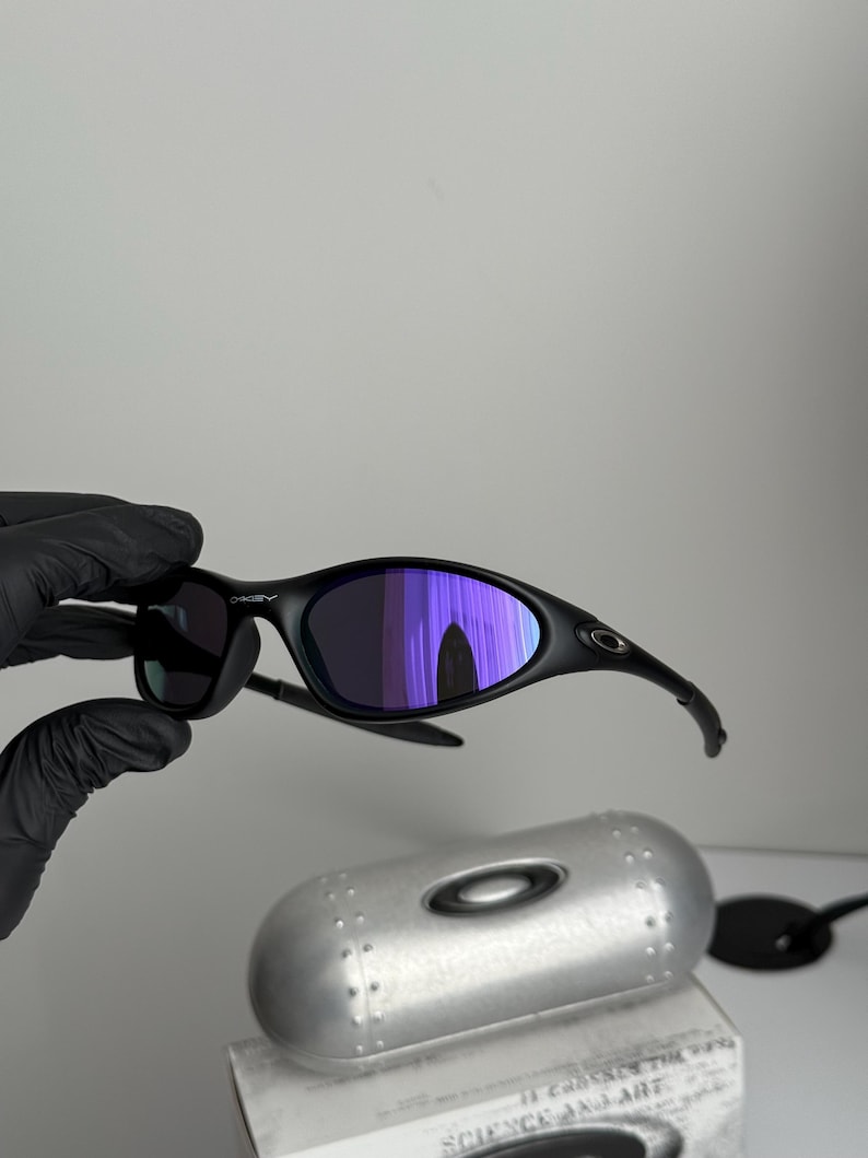 May include: Black Oakley sunglasses with purple mirrored lenses. The sunglasses are held by a gloved hand. A silver case and a box with the text "SCIENCE AND ART" are in the background.