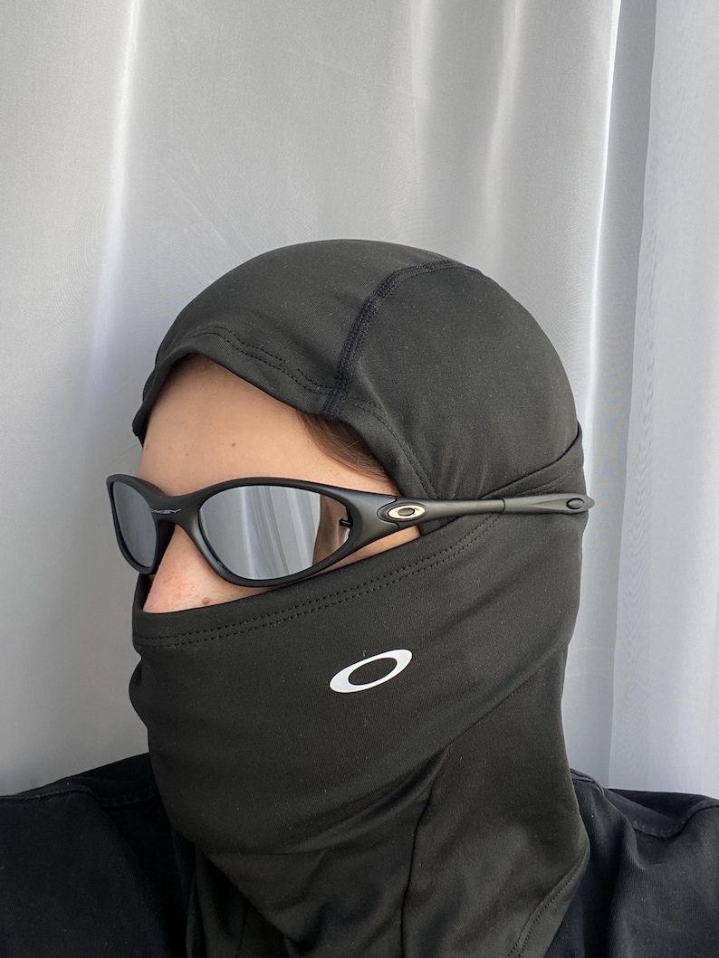 Puede incluir: Pasamonta&ntilde;as negro con un logotipo blanco y gafas de sol negras. Las gafas de sol tienen montura negra y lentes espejadas. El pasamonta&ntilde;as cubre la cabeza y el cuello.