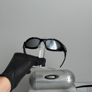 May include: Black sunglasses with reflective lenses are displayed on a clear stand. A silver case with rivets and the Oakley logo sits below the glasses. The box below the case has the text "SCIENCE AND ART".