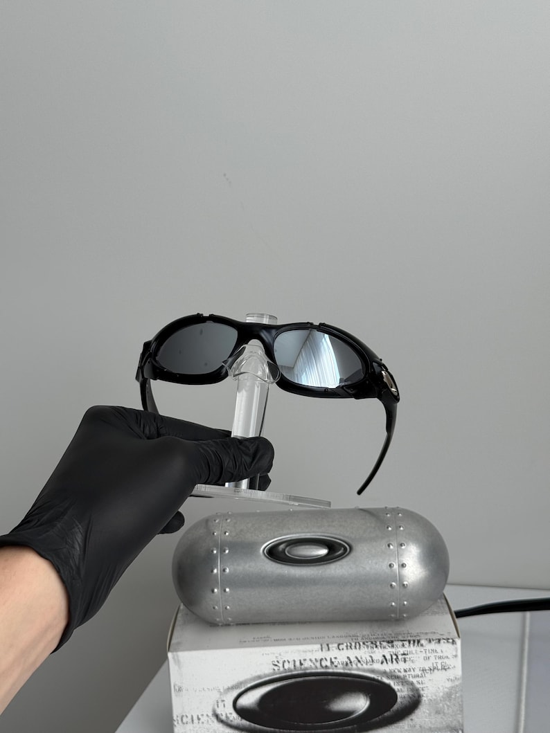 May include: Black sunglasses with reflective lenses are displayed on a clear stand. A silver case with rivets and the Oakley logo sits below the glasses. The box below the case has the text "SCIENCE AND ART".