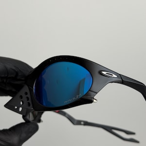 May include: Black sunglasses with blue mirrored lenses. The sunglasses have a sleek, modern design with a matte finish. The lenses are curved and feature the word "PRIZM". The arms are thin and curved.