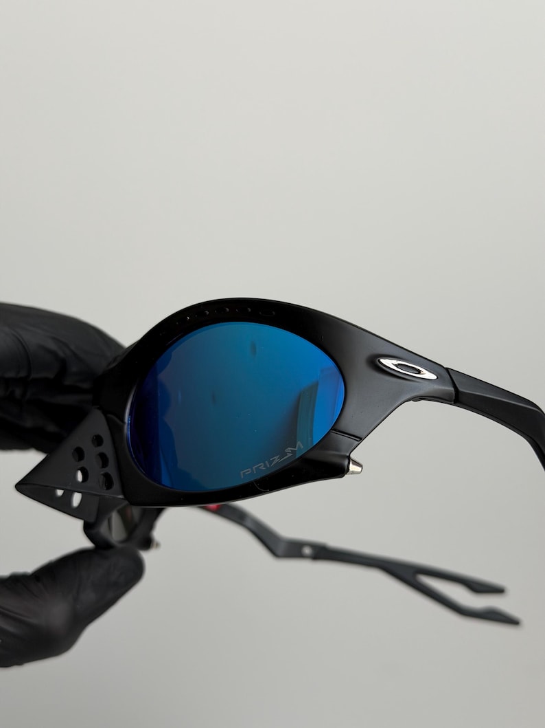 May include: Black sunglasses with blue mirrored lenses. The sunglasses have a sleek, modern design with a matte finish. The lenses are curved and feature the word "PRIZM". The arms are thin and curved.