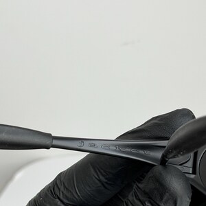 May include: Black sunglasses with the word "jacket" etched on the arm. The sunglasses are held by a gloved hand against a white background. The frame is a matte black.