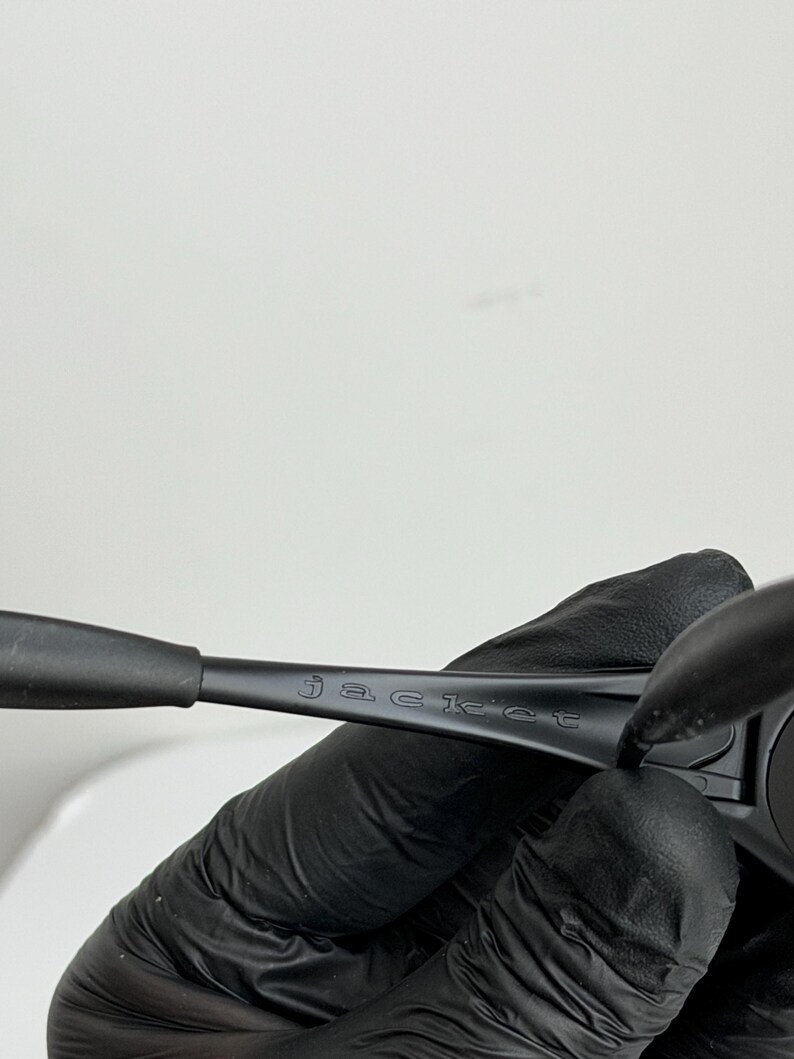 May include: Black sunglasses with the word "jacket" etched on the arm. The sunglasses are held by a gloved hand against a white background. The frame is a matte black.