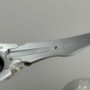 May include: Close-up of a silver-colored sunglasses arm. The text "MADE IN U.S.A." is engraved on the arm. The sunglasses arm is made of a metallic material and has a sleek, modern design. The background is a solid gray.